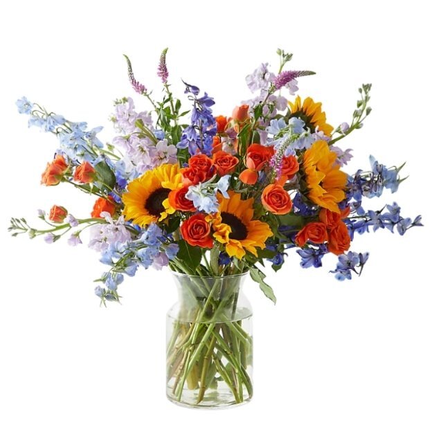 Bright floral arrangement featuring sunflowers, orange and red roses, and blue accent flowers in a clear glass vase.