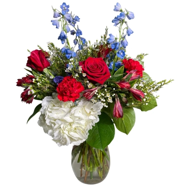 Vibrant floral arrangement featuring red roses, blue flowers, white hydrangea, and seasonal accents in a round glass vase.